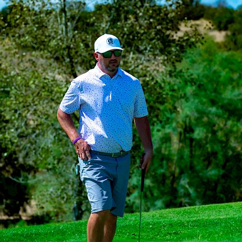 A man in a white hat and sunglasses is standing on a golf course, holding a putter, with trees in the background and a bright, clear sky.