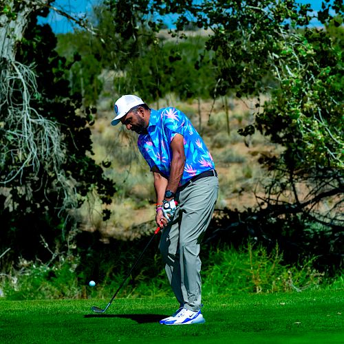 A person is playing golf on a bright, sunny day, wearing a colorful shirt, gray pants, and a white cap, surrounded by trees and green grass.