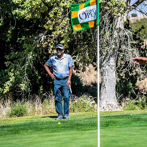 A golfer stands near a green, observing the position of their ball next to a hole marked by an OPEI flag, with trees in the background.