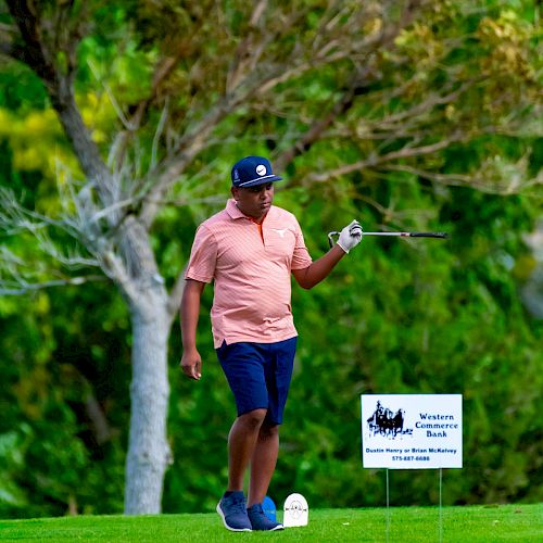 A person is standing on a golf course holding a golf club, with a tree in the background and a sign next to them.