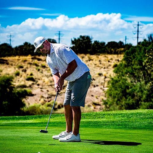 A person wearing a white shirt and gray shorts is putting on a golf course, with a scenic background of trees and blue sky.