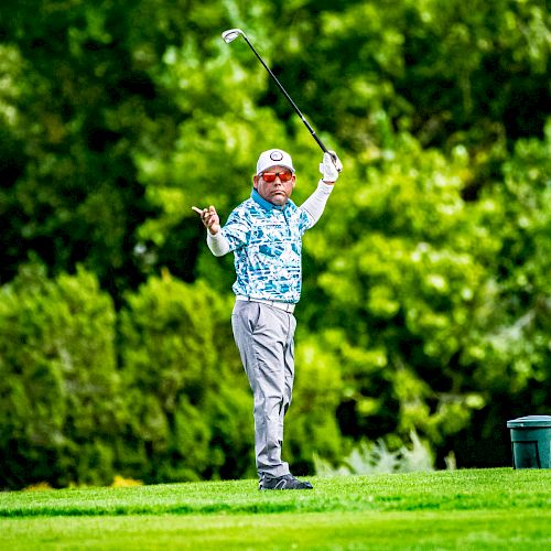 A golfer mid-swing on a golf course, surrounded by lush greenery and trees in the background, wearing a floral shirt and a cap, with a bucket nearby.