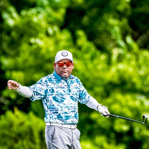 A man wearing a patterned shirt, cap, and sunglasses holds a golf club, standing against a backdrop of lush green foliage.