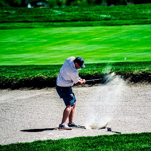 A person is hitting a golf ball out of a sand bunker on a golf course, with grass and trees in the background.