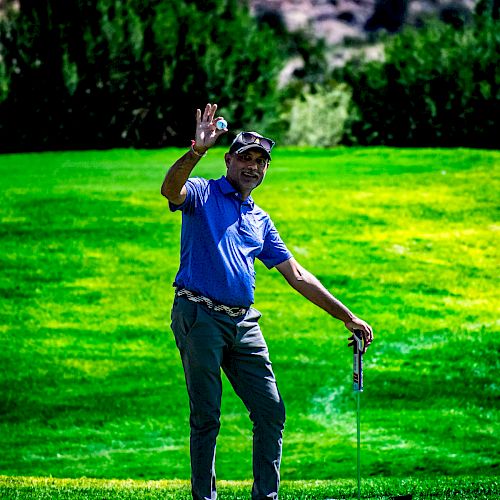 A person standing on a golf course waves while holding a golf club. Lush green grass surrounds them with trees and hills in the background.