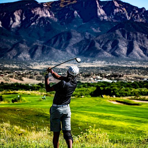 A golfer is swinging a club on a green fairway with towering mountains in the background under a bright blue sky.