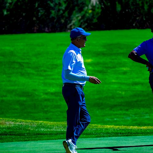 Two individuals stand on a vibrant golf course, with one person in the foreground walking and the other partially visible.