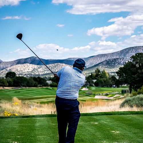 A golfer is mid-swing on a lush green course, surrounded by trees and mountains under a partly cloudy blue sky.