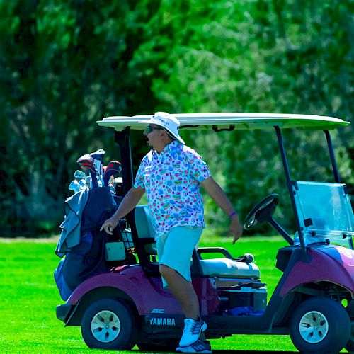 A person wearing a floral shirt and hat stands by a golf cart on a green golf course, with trees in the background.