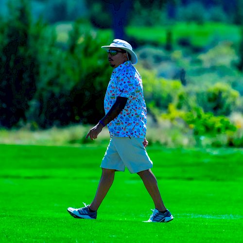 A person wearing a hat, floral shirt, shorts, and sneakers is walking on a grassy field with trees and shrubs in the background.