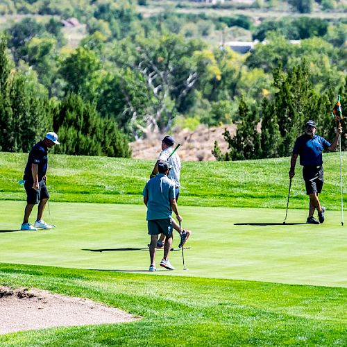 Four individuals are on a golf course putting green, engaged in a game of golf during a sunny day with lush greenery in the background.