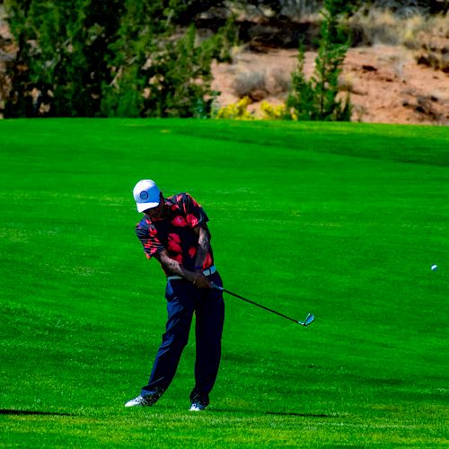 A golfer in a plaid shirt and dark pants is swinging a club on a vibrant green golf course with trees and a dirt area in the background.