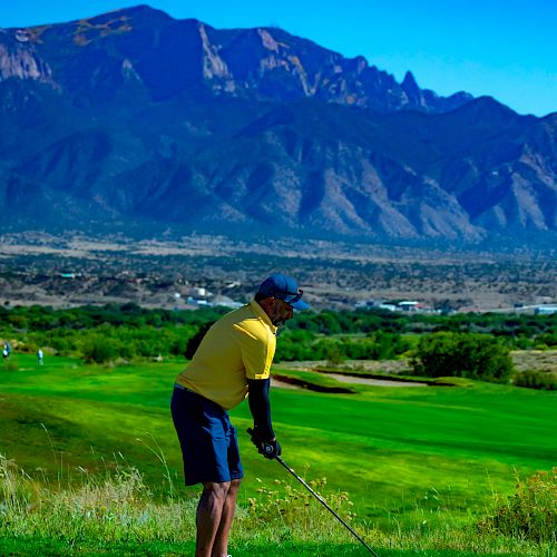 A person is golfing on a lush, green course with mountains in the background under a bright blue sky, ready to swing the golf club.