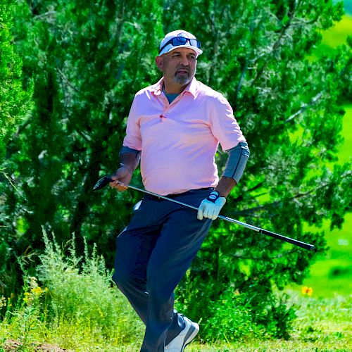 A person wearing a pink shirt, blue pants, and a cap is holding a golf club while standing on a green field with trees in the background.