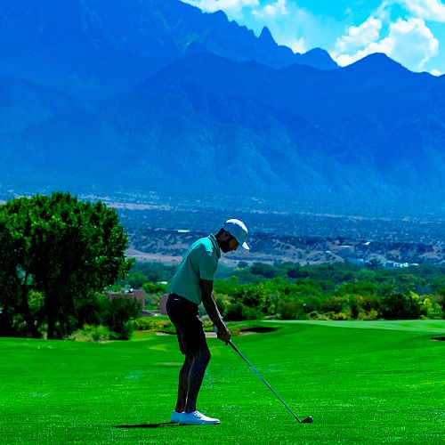 A golfer prepares to swing on a lush green course with a scenic mountain backdrop under a partly cloudy sky.