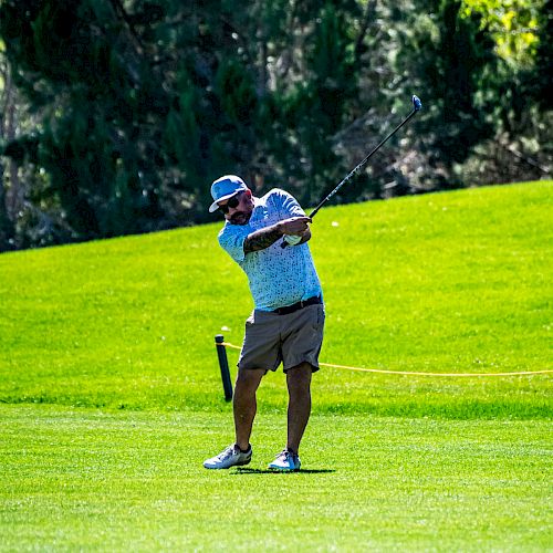 A person mid-swing on a golf course, wearing a cap and golf attire, with a background of green grass and trees.