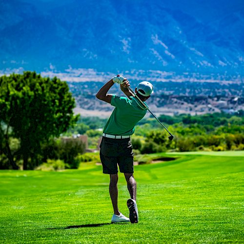 A golfer is mid-swing on a lush green golf course with mountains in the background, under a bright blue sky and scattered clouds.