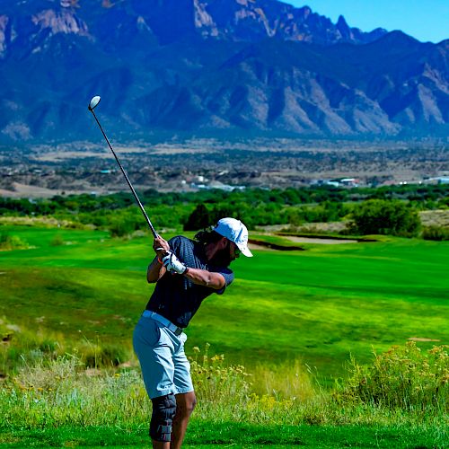 A golfer mid-swing on a lush green course with a mountainous backdrop under a clear blue sky.