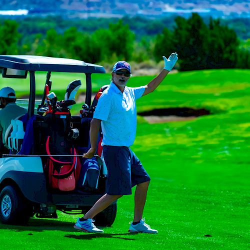 A person is standing by a golf cart on a golf course, waving with a smile, surrounded by green grass and a scenic background.