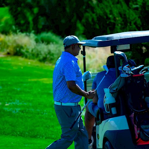 A man in golf attire stands beside a golf cart loaded with clubs on a green golf course, surrounded by trees and shrubs.