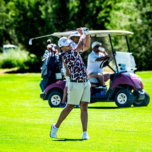 A person is playing golf, swinging a club on a green field, with a golf cart and another person in the background.