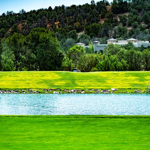 The image shows a lush green lawn in the foreground, a small pond or water feature, and distant greenery, with buildings partially visible behind trees.