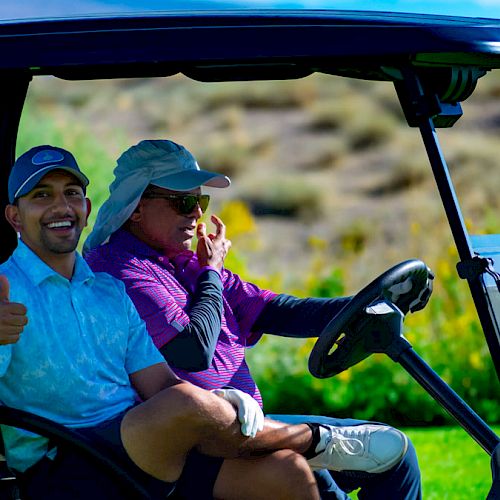 Two men are sitting in a golf cart, with one giving a thumbs-up and smiling while the other is talking on a phone.