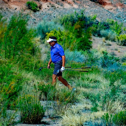 A person wearing a blue shirt and a hat walks through a grassy, dry landscape carrying a stick. The background features rocky terrain and shrubs.