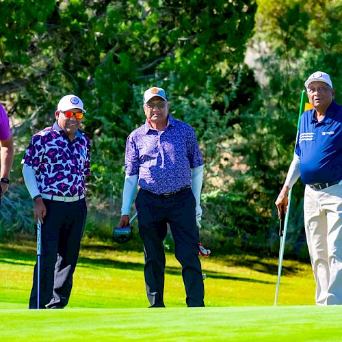 Four men are standing on a golf course, each holding a golf club, with greenery in the background.