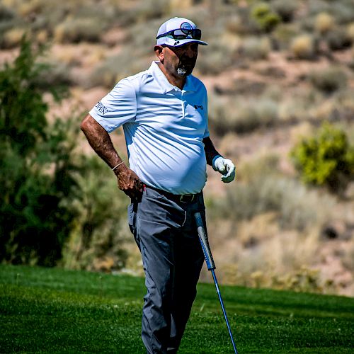 A man in a white polo and gray pants stands on a golf course, holding a golf club with one hand on his hip. The background is a dry, hilly landscape.