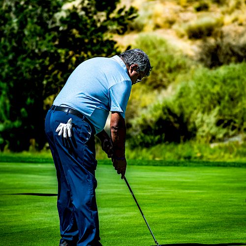 A man is playing golf on a lush green course, concentrating on his shot with a putter, surrounded by trees and hills in the background.