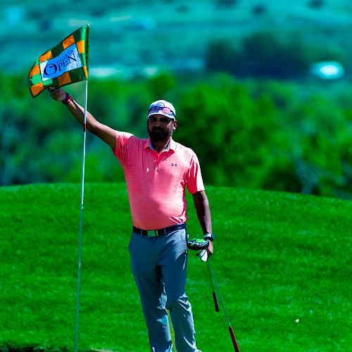 A golfer in a pink shirt and gray pants holds a flagstick on a putting green with a golf club in his other hand, against a green landscape.