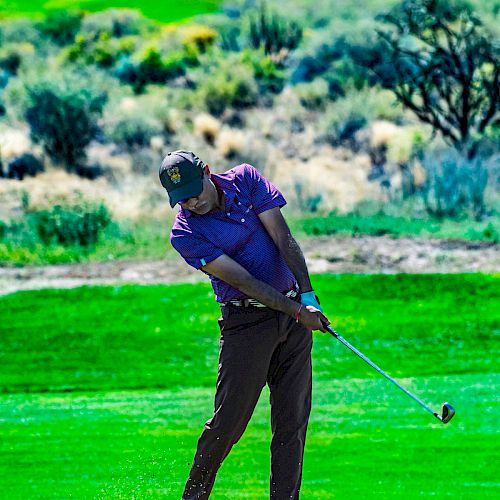 A person is playing golf on a lush green course, mid-swing with a golf club, wearing a purple shirt and brown pants, with greenery in the background.