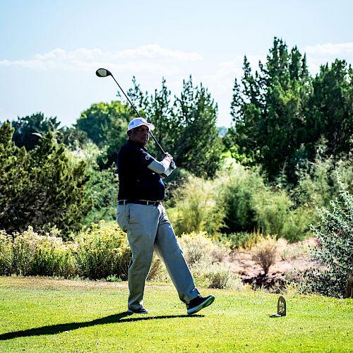 A person is playing golf, swinging a club on a lush green course with trees and shrubs in the background under a clear sky.