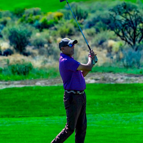 A person dressed in a purple shirt and dark pants is playing golf, swinging a golf club on a lush green golf course with trees in the background.