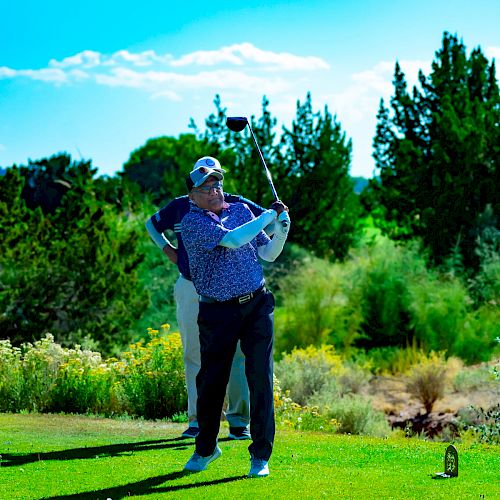 A man is playing golf on a sunny day, in the motion of swinging his club, surrounded by green trees and grass. Another person is in the background.