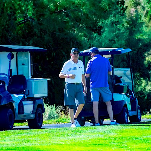 The image shows two men standing near their golf carts, conversing on a golf course, with green grass and trees visible in the background.