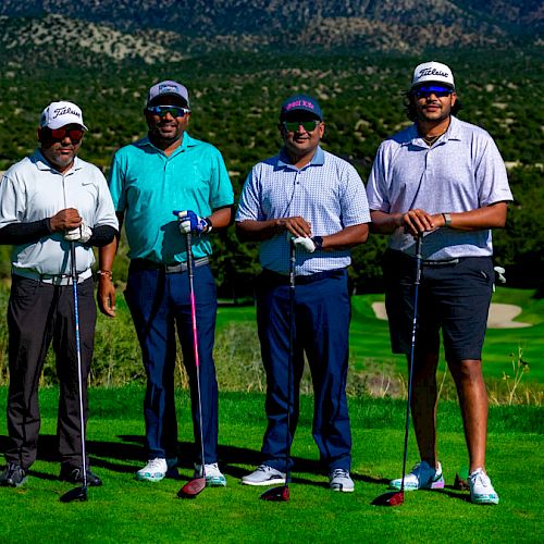 Four men stand on a golf course holding clubs, with green fairways and hills in the background. They are wearing golf attire and caps.