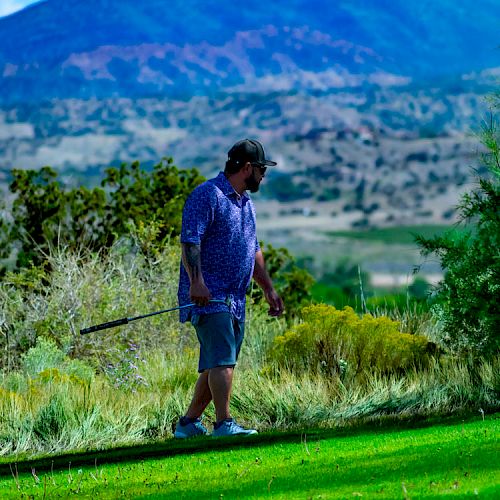 A person holding a fishing rod is walking on a grassy area in a scenic outdoor setting with mountains and trees in the background.