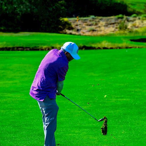 A person dressed in a purple shirt and gray pants is hitting a golf ball on a lush green golf course, with trees and blue sky in the background.