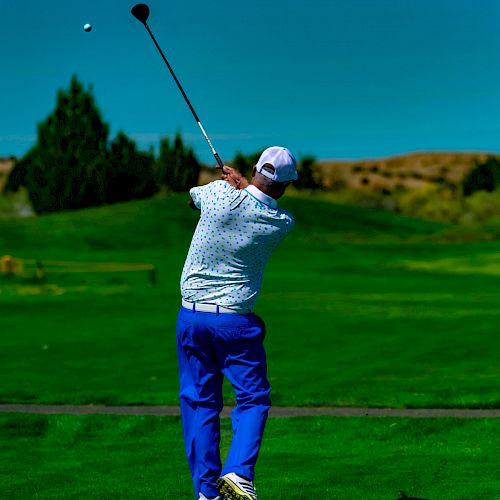 A golfer wearing a white cap and shirt with blue pants is swinging a club on a green golf course under a clear blue sky.