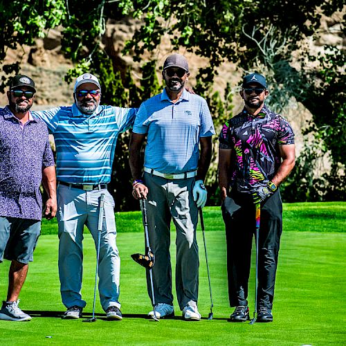 Four people are standing on a golf course; they are dressed in golf attire and holding golf clubs, posing for a group photo.