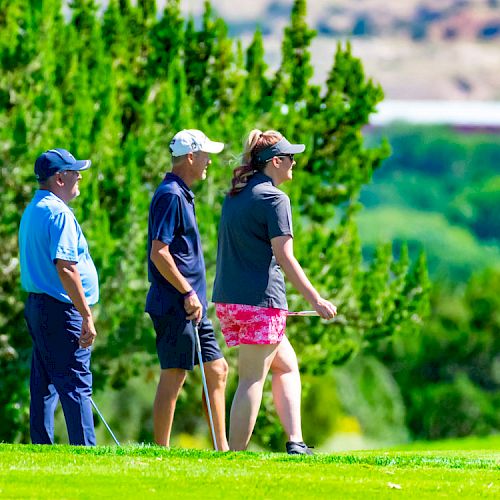 Three people stand on a lush green golf course, with trees and a clear sky in the background.