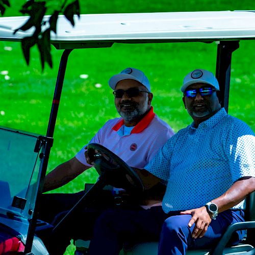 Two people are sitting in a golf cart on a grassy course, wearing caps and sunglasses, with trees in the background.