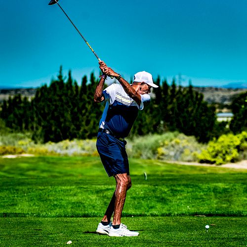 A person is in mid-swing on a sunny golf course, wearing a white hat, navy shirt, and shorts, surrounded by greenery and a clear blue sky.