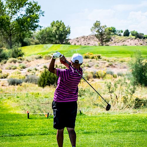 A golfer in a pink striped shirt and black shorts is swinging a club on a green golf course with trees and hills in the background.