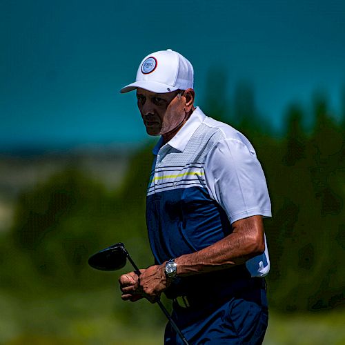 A person wearing a cap and golf attire is holding a golf club on a golf course, with trees and a clear sky in the background.