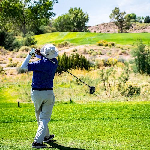 A person in a blue shirt and white pants is playing golf on a green course, captured in mid-swing under a sunny sky with trees in the background.