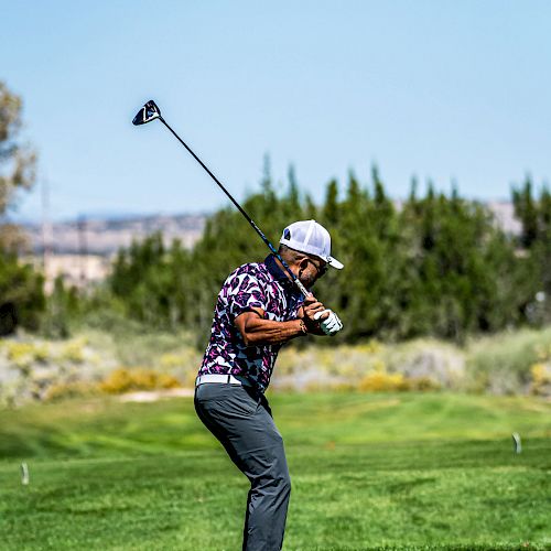 A person on a golf course is in mid-swing, wearing a cap, patterned shirt, and dark pants. Trees and a clear sky are visible in the background.