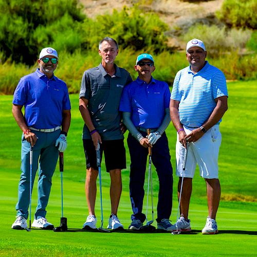 Four men standing on a golf course, each holding a golf club, with greenery and a hill in the background.